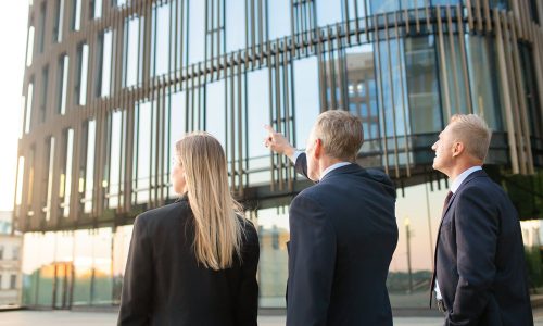 Group of business partners in formal suits pointing at office building, meeting outdoors, discussing real property. Back view. Commercial real estate concept